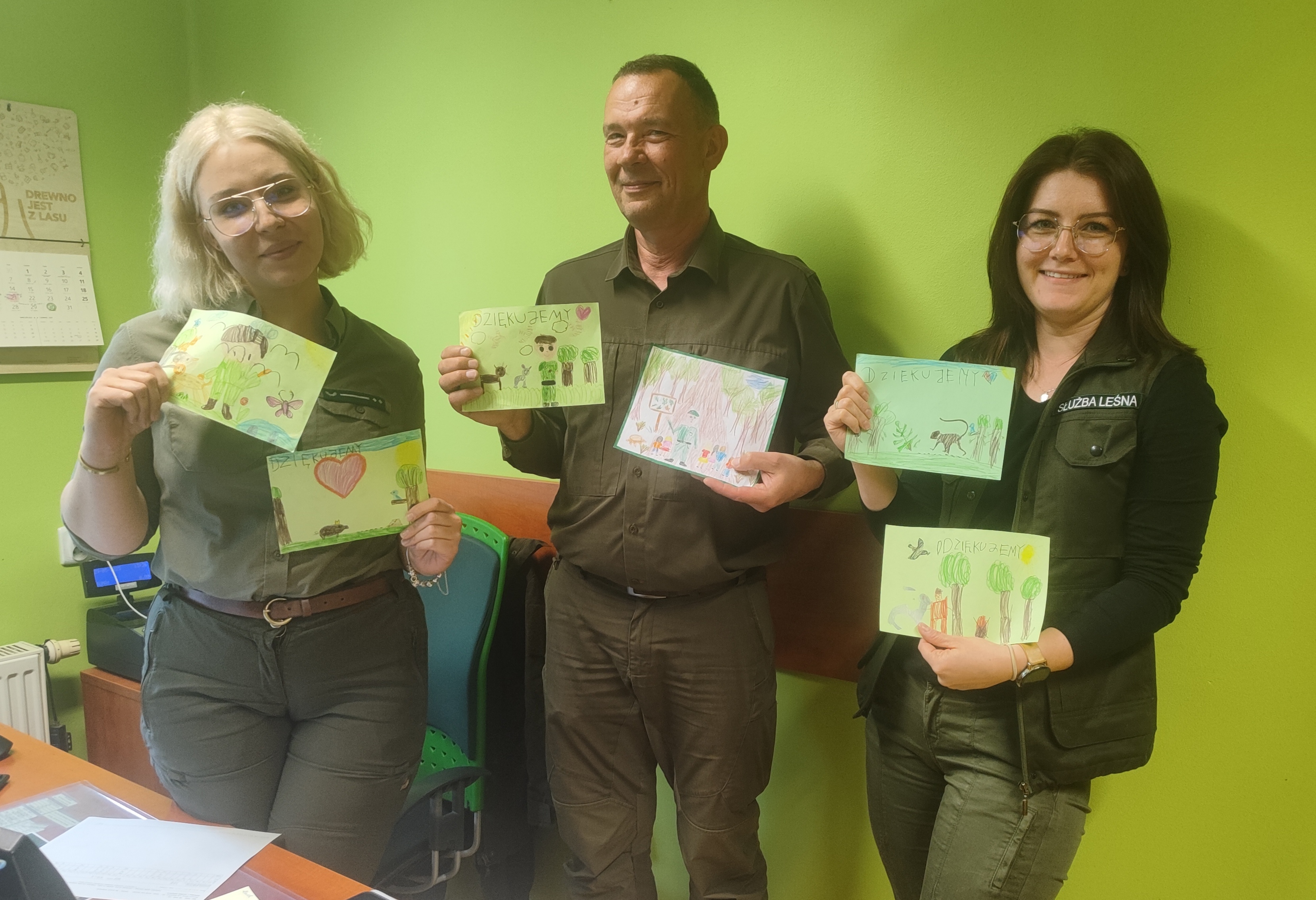 Dwie Moniki i Kuba dziękują za wspaniałą niespodziankę! Zdjęcie przedstawia leśników - dwie kobiety i mężczyznę, którzy uśmiechnięci stoją w pomieszczeniu, trzymając w rękach rysunki dzieci. (fot. archiwum nadleśnictwa, M.Prętkowska)