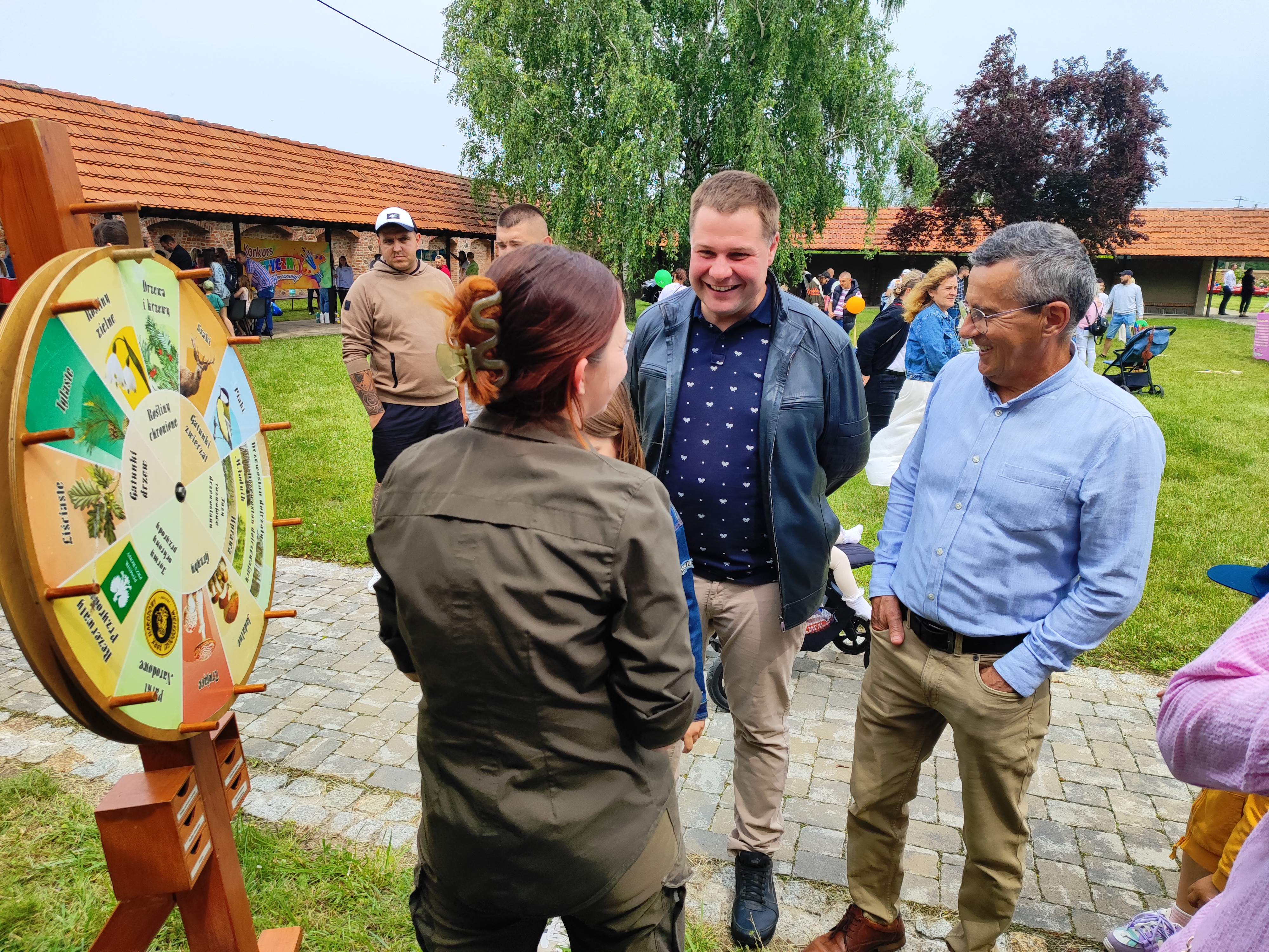 Zdjęcie przedstawia uśmiechniętych ludzi stojących przy stoisku nadleśnictwa, rozmawiających z kobietą - leśnikiem, w tle wiaty z pomarańczowym zadaszeniem i dwa duże drzewa. (fot. archiwum nadleśnictwa, M. Prętkowska) 