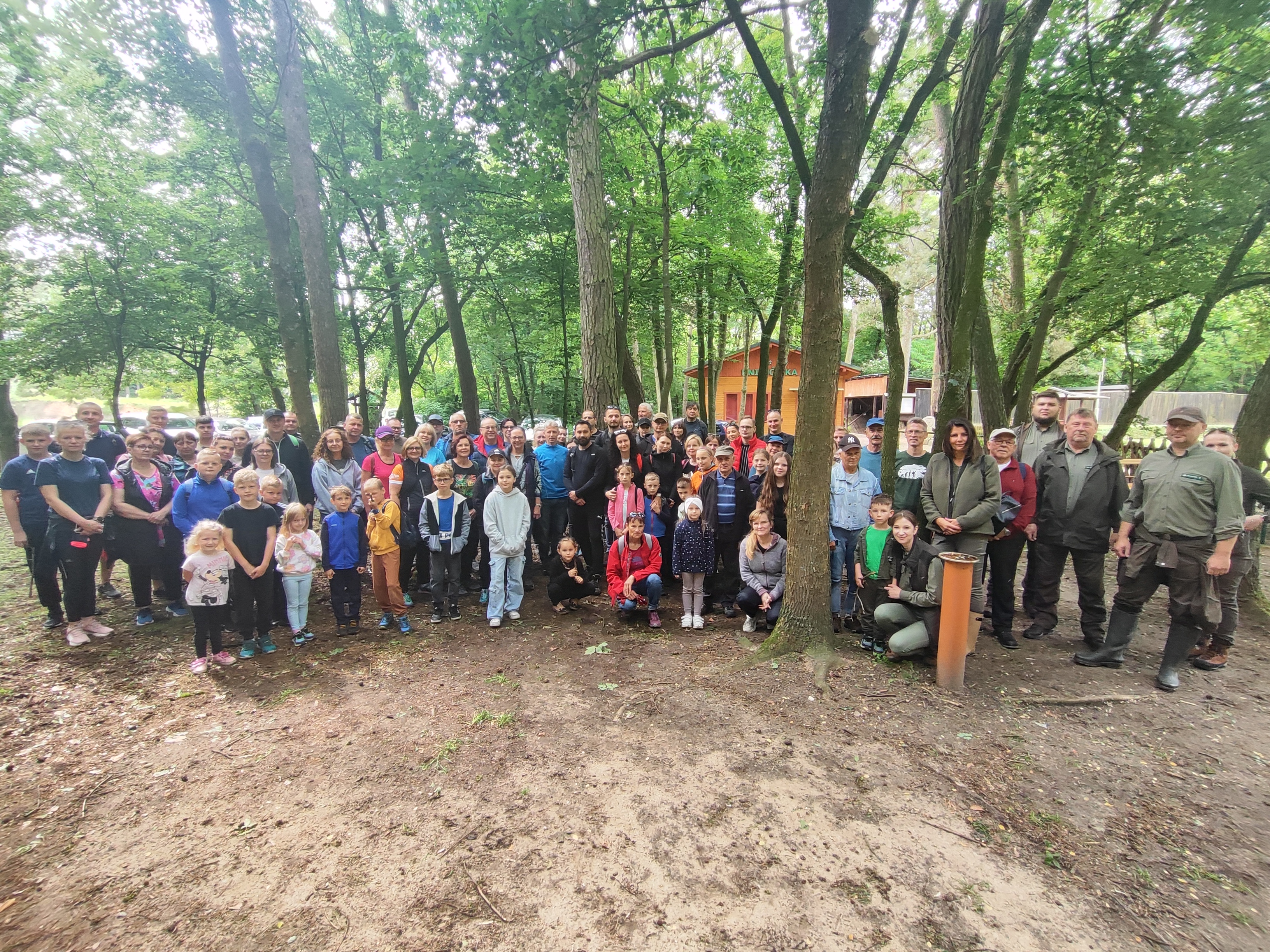 Zdjęcie przedstawia uczestników spaceru przyrodniczego podczas zbiórki, w tle drzewa i drewniany budynek. (fot. Sołectwo Piaski)