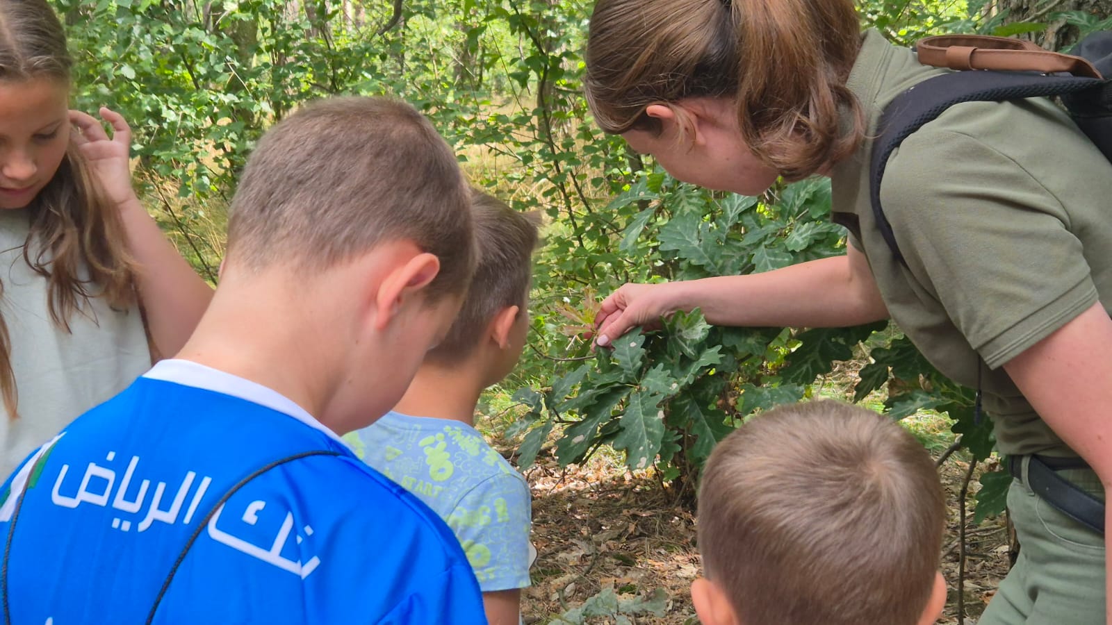Zdjęcie przedstawia kobietę leśnika  i grupę dzieci stojących przy drzewie i obserwujących liście. (fot. CKiB Piaski)
